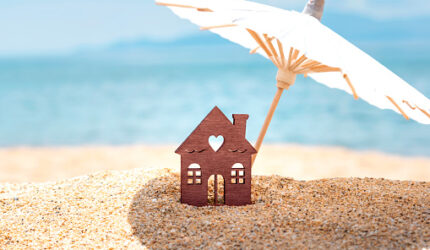 Miniature house and umbrella on beach, blue sea and sky on blurred background. Real estate, sale or property investment concept. Symbol of dream home for family. Copy space