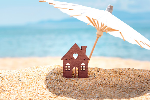 Miniature house and umbrella on beach, blue sea and sky on blurred background. Real estate, sale or property investment concept. Symbol of dream home for family. Copy space